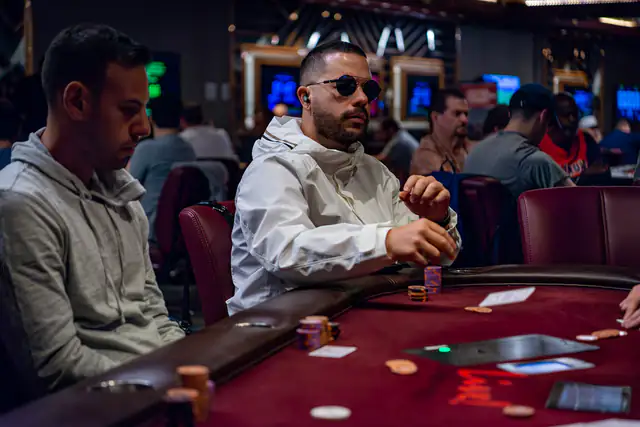 Players seated around a red poker table inside a busy card room